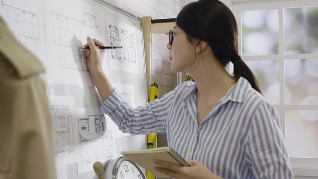 slow action focused casual asian female architect working drawing board with pen and notebook in hand. woman interior designer wearing glasses stand in office. Floor plan busy concentration concept