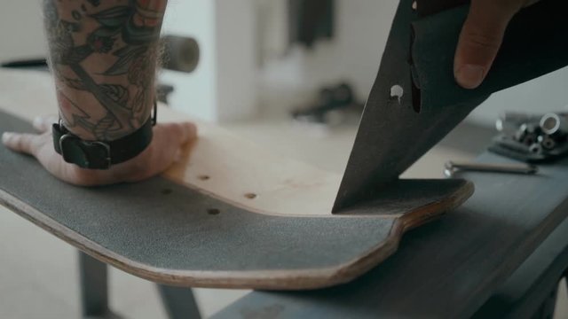 Millennial skateboarder or artisan boards maker recycles his old deck, regrips it using razor blade. Removes grip tape by pulling it with hands. Small business owner or hobby