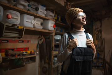 Portrait of smiling young builder girl standing in old workshop room, wearing construction glasses.