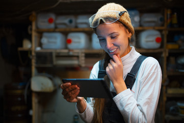Portrait of young builder girl standing in workshop use smartphone, wearing construction glasses.