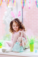 child girl with pink rabbit ears with an Easter bunny in her hands sits on the windowsill against the window.