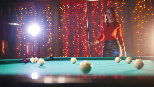 Billiards club. Concentrated ginger woman in glasses aiming and hitting the ball