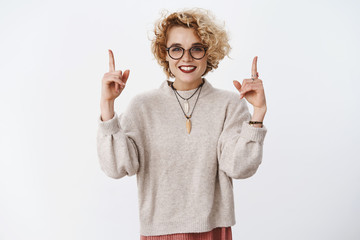 Studio shot of stylish queer girl with short blond hair lipstick and glasses pointing up with...