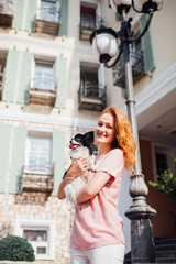 The theme is the friendship of man and animal. Beautiful young red hair Caucasian woman holding a pet dog Chihuahua breed near a house building in the summer in sunny weather