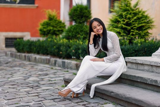 Close up portrait of beautiful asian girl dressed in Ao Dai white dress sitting on the stairs in courtyard.