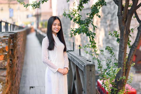 Close up portrait of beautiful asian girl dressed in Ao Dai white dress stands near the tree on wooden bridge.