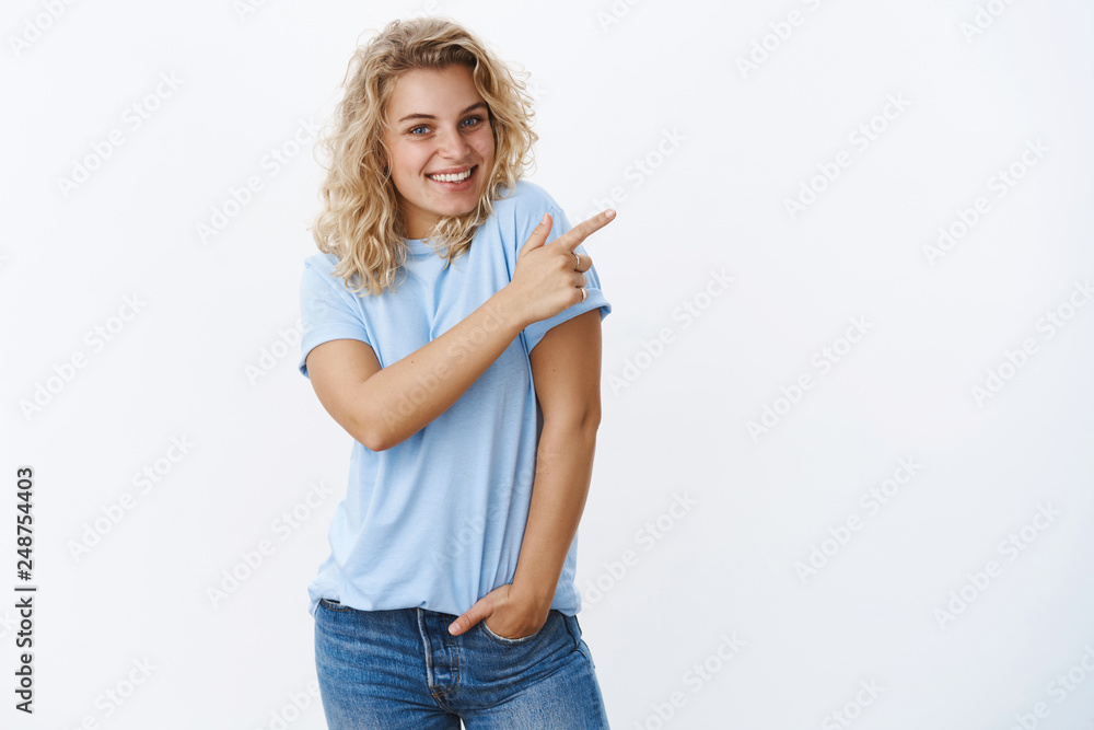 Wall mural Charming enthusiastic and joyful blond woman with blue eyes in t-shirt point at upper right corner and smiling happily as pleased, showing awesome product with positive impression after using it - Wall murals