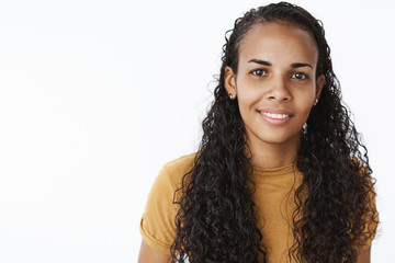 Cute and sincere young charismatic african-american female with long curly natural hair smiling joyfully at camera with white teeth gazing at camera optimistic and satisfied against gray wall
