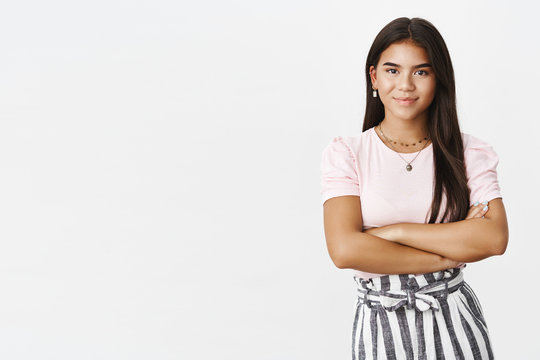 Studio shot of assertive and self-assured good-looking young adolescent girl in skirt and blouse holding hands crossed over chest in confident and ready pose, smiling friendly at camera