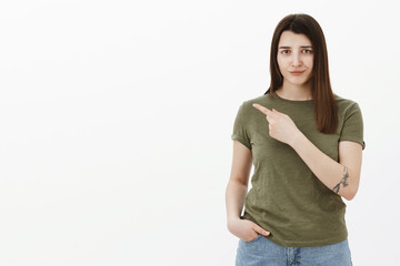 Shy, unsure insecure cute brunette girl with tattto smirking and frowning with sad unconfident expression as pointing at upper left corner or behind asking take look, hesitant and timid over gray wall