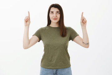 Confident and assertive attractive female student with tattoo in olive t-shirt looking self-assured up as pointing upwards and smiling delighted, ready for action, posing over white background