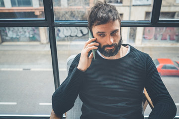 Cheerful caucasian man in black pullover using mobile phone application for mobile blogging. Confident young hipster guy read news from social network at modern loft.Blurred background