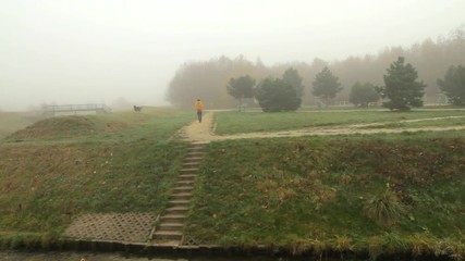 A woman with her dog are taking their morning walk through a foggy city park.