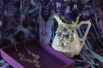 white porcelain pitcher with purple flowers next to a purple box with a pretty necklace with a...
