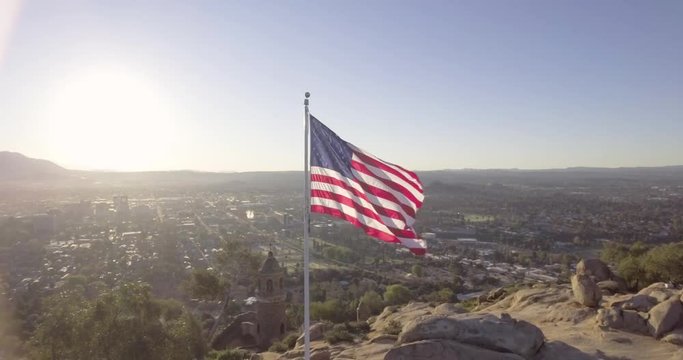 Drone aerial of an American flag flying on a mountain top overlooking a city at sunrise with light flares