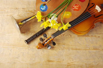 Yellow daffodils, violin, trumpet, art palette and attributes of arts on a wooden table.