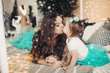 Mother kissing her daughter, lying on bed. Beautiful woman having long curly chestnut hair. Little...