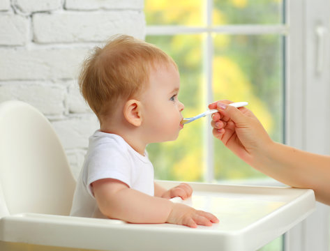 Baby 7 months in a child seat in front of the window eats food from a spoon