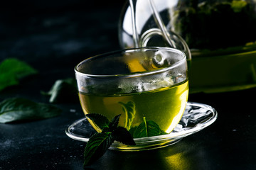 Hot chinese green tea with mint, with splash pouring from the kettle into the cup, steam rises, dark background, selective focus
