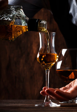The bartender pours the grappa or liquor in small shot glass on the old bar counter. Vintage wooden background in pub or bar, night mood. Place for text, toning, selective focus