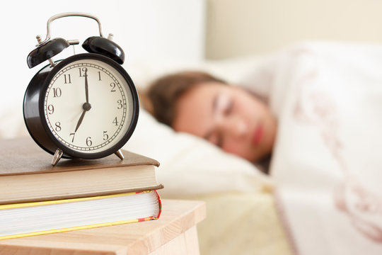 Teenager girl sleeping in a white bed. Alarm clock in the foreground on a pile of books