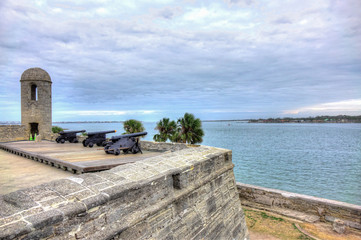 Three Spanish colonial cannons mounted on top of an old Spanish fort, overseeing the ocean, a cloudy sky and palm trees. Shot in Castillo de San Marcos, St Augustine, Florida, USA.