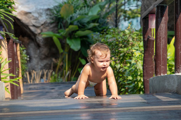 Fototapeta premium Funny baby learns to crawl. dynamic movement. Child crawls along the dark brown wooden walkway of the bridge at sunset. Baby learn crawl on the background of greenery and a wooden bridge. There is a