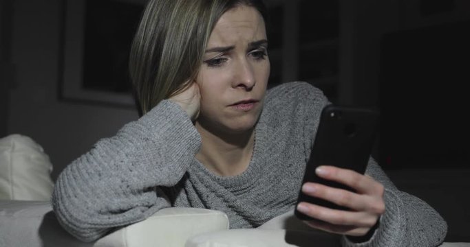 A woman sits in a dark room at home while using a smartphone and looks concerned and worried
