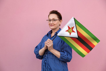 Zimbabwe flag. Woman holding Zimbabwe flag. Nice portrait of middle aged lady 40 50 years old holding a large flag over pink wall background on the street outdoors. - 248806673