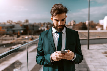 Portrait of successful Caucasian businessman in formal wear using smart phone on rooftop. In...
