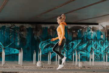 Young beautiful blonde Caucasian woman in sportswear and with ponytail jumping under the bridge. Full length.
