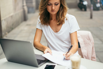 Pretty female freelancer making notes and diagrams in papers while having a distant work at laptop, young girl watching an online video lesson and studying using notebook and papers  sitting in a cafe