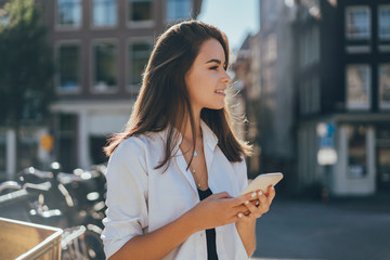 Pretty smiling happy girl holding mobile phone in her hands and browsing internet while waiting to her friend standing on the street, typing in messenger and scrolling web pages