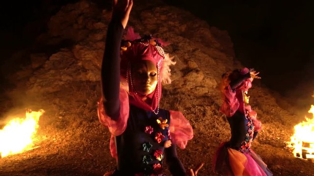 dancers girls in purple and pink dresses and gold masks are holding one hand on the belt, and the second over his head. At night with bonfires in the background