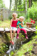 Happy smiling children sitting on the bridge at the creek embracing one another in a country house garden. Kids playing outdoors in summer, vertical