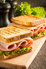 Close-up of two sandwiches with bacon, salami, prosciutto and fresh vegetables on rustic wooden cutting board. Club sandwich concept.