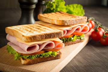 Close-up of two sandwiches with bacon, salami, prosciutto and fresh vegetables on rustic wooden cutting board. Club sandwich concept.