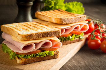 Close-up of two sandwiches with bacon, salami, prosciutto and fresh vegetables on rustic wooden cutting board. Club sandwich concept.