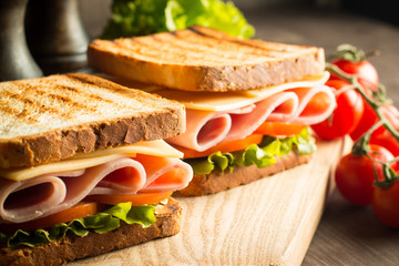 Close-up of two sandwiches with bacon, salami, prosciutto and fresh vegetables on rustic wooden cutting board. Club sandwich concept.