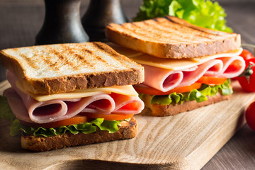 Close-up of two sandwiches with bacon, salami, prosciutto and fresh vegetables on rustic wooden cutting board. Club sandwich concept.