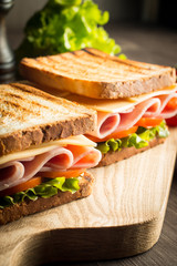 Close-up of two sandwiches with bacon, salami, prosciutto and fresh vegetables on rustic wooden cutting board. Club sandwich concept.