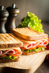 Close-up of two sandwiches with bacon, salami, prosciutto and fresh vegetables on rustic wooden cutting board. Club sandwich concept.