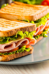 Close-up of two sandwiches with bacon, salami, prosciutto and fresh vegetables on rustic wooden cutting board. Club sandwich concept.