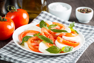 Photo of Caprese Salad with tomatoes, basil, mozzarella, olives and olive oil on wooden background. Italian traditional caprese salad ingredients. Mediterranean, organic and natural food concept.