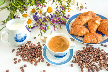 Summer breakfast concept: cup of coffee with foam, hot croissants, jug of milk, coffee beans and bunch of daisies on white wooden background