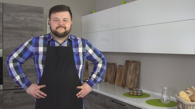 Tight kind man in an apron with a beard is standing in the kitchen and looking at the camera, copy space, 4K