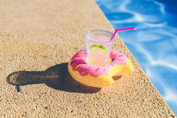 Fototapeta na wymiar Tasty refreshing cocktail on on inflatable donut plastic toy on edge of swimming pool. Ripple Water in swimming pool with sunny reflection