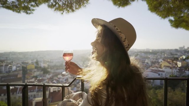 Side view of beautiful young female smiling and looking away while drinking good wine on terrace of outdoor cafe on sunny day