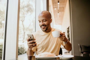 Happy cheerful African hipster holding mug, drinking fresh cappuccino, browsing internet and checking newsfeed on social media.Man using cell phone during coffee break at modern cafe