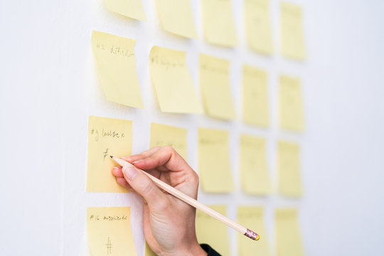 The hand of a woman writing on post it notes on a white wall with a pencil.